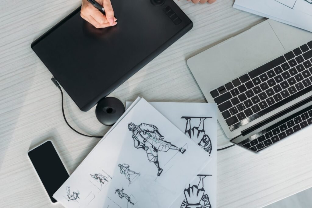 Vista superior de una mesa de trabajo que muestra una mano usando una tableta gráfica conectada a una laptop. También hay un teléfono móvil, un cuaderno con ilustraciones de personajes y un lápiz óptico.